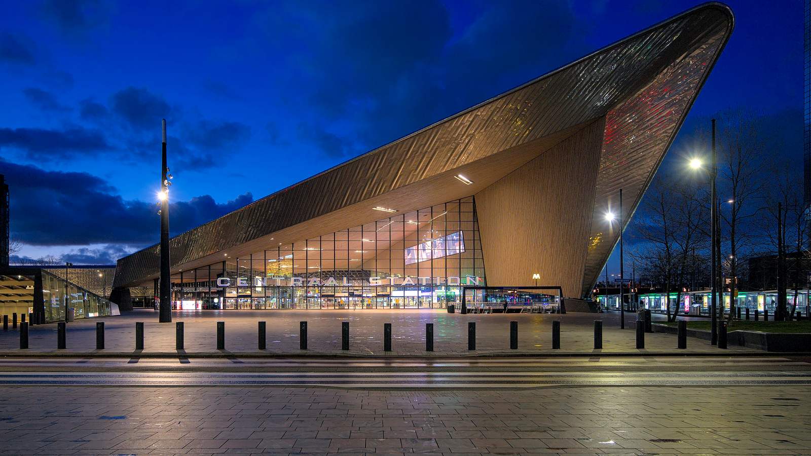 De stationshal van Rotterdam Centraal met het puntige, metalen dak en de verlichte glazen gevel bij schemering