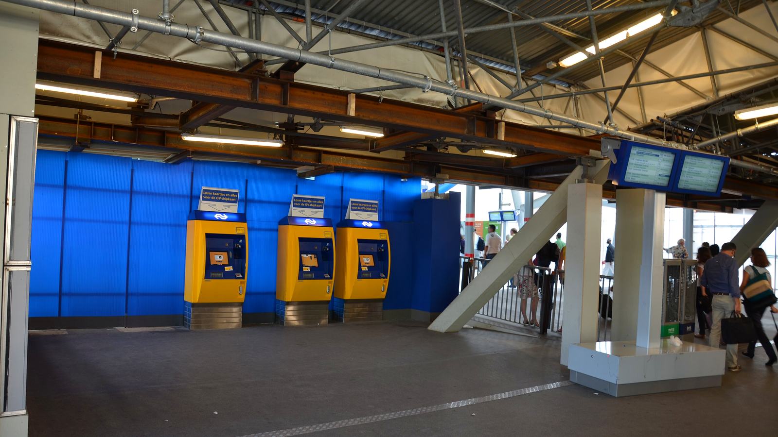 Drie gele kaartautomaten van NS tegen een blauwe wand op station Utrecht Centraal, met reizigers op weg naar de perrons