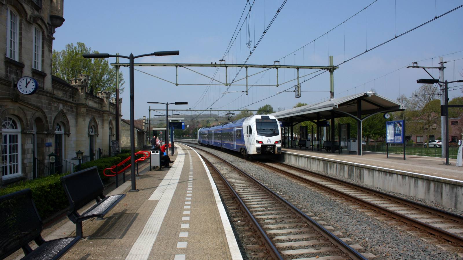 Wit-blauwe FLIRT-trein van Arriva rijdt station Valkenburg binnen, links het monumentale stationsgebouw met klok en een perron met bankjes